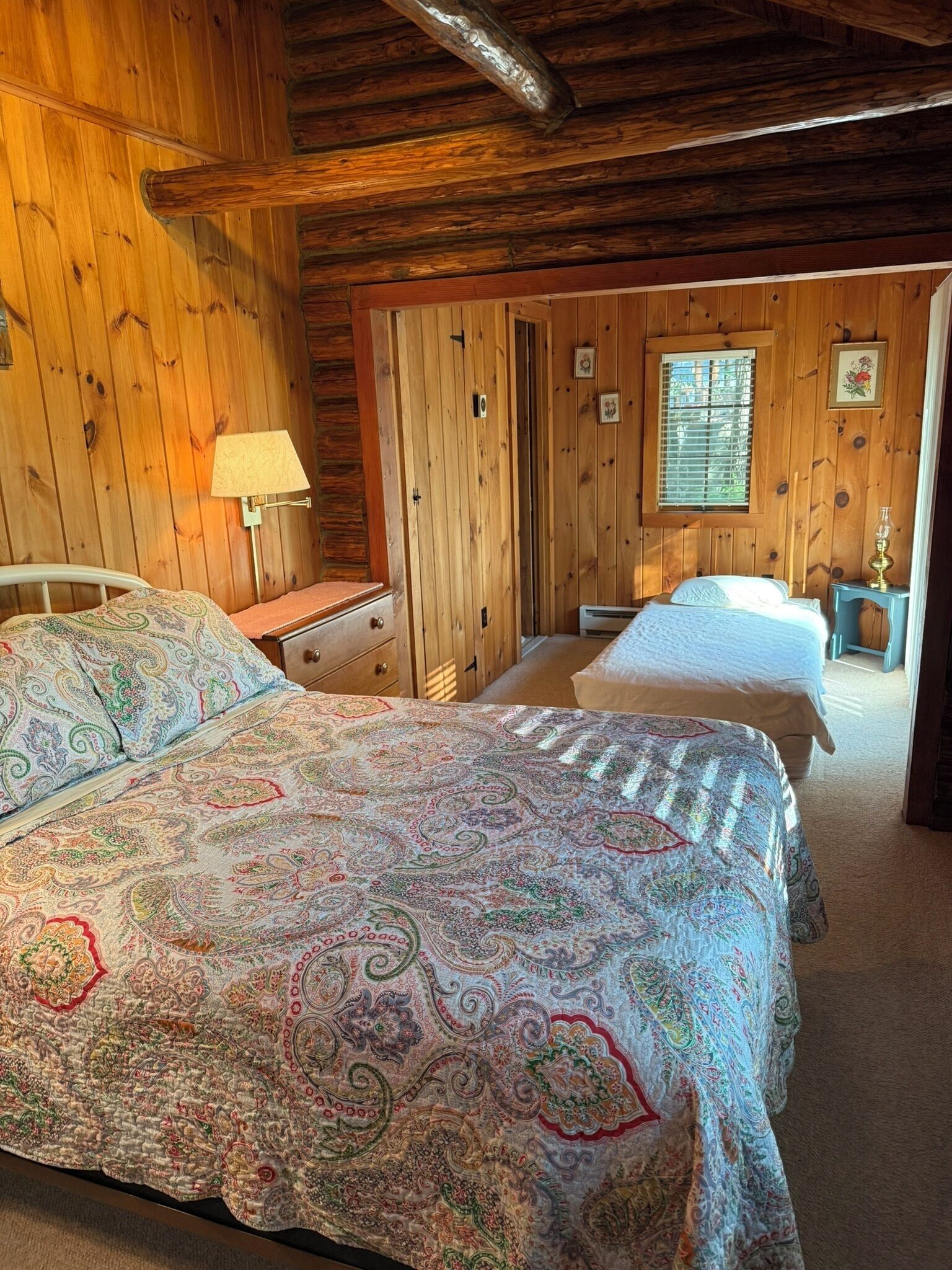 Un dormitorio de cabaña de madera con dos camas, una mesita de noche, una lámpara y una ventana con persianas.