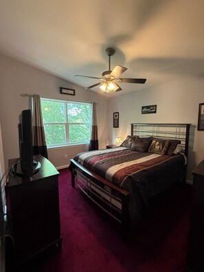 A bedroom with a bed, a ceiling fan, a window with curtains, and a television on a stand. - Rocky Road Chalet - Masthope (Lackawaxen)