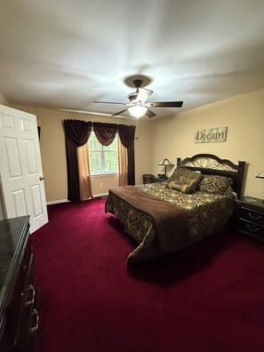 A bedroom with a bed, a ceiling fan, a window with curtains, a nightstand, and a wall with a framed picture.