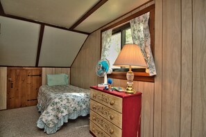 Quarto com cama, cômoda, luminária, ventilador e janela com cortinas.