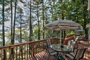 A deck with a glass table and chairs, surrounded by a wooden railing, overlooking a lake with trees. - Sunnyview Wallenpaupack Lakefront Chalet Spectacular Sunsets  private dock (Paupack)