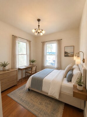 Un dormitorio con cama, mesita de noche, escritorio, silla y vistas al exterior.