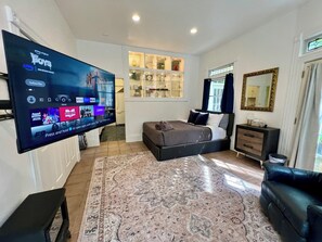 A modern bedroom with a large flat-screen TV, a bed with dark bedding, a bedside table, a dresser, and a window with curtains. - Charming 4-Bed Sausalito Home with Bay Views & Garden Patio near San Francisco (Sausalito)