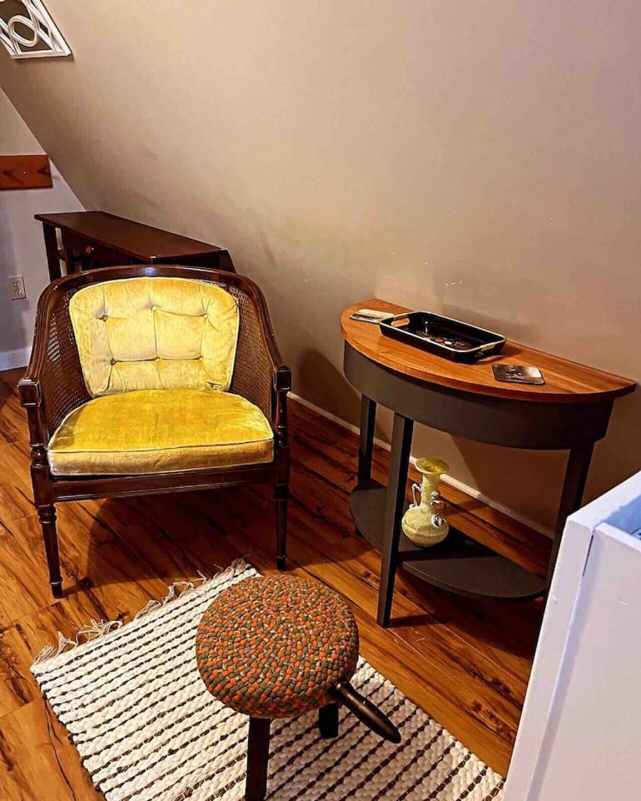 Een kamer met een houten vloer, een geel bekleden stoel, een klein rond krukje, een ronde houten tafel met een zwart object erop, en een witte muur.
