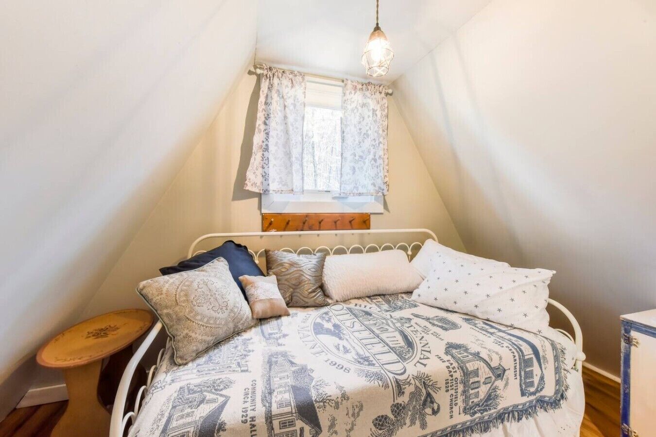 Une chambre avec un plafond en pente, un lit simple avec une literie à motifs, une petite table et une fenêtre avec un rideau.