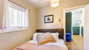 A bedroom with a bed, a window with curtains, a framed picture on the wall, and a view into the kitchen.