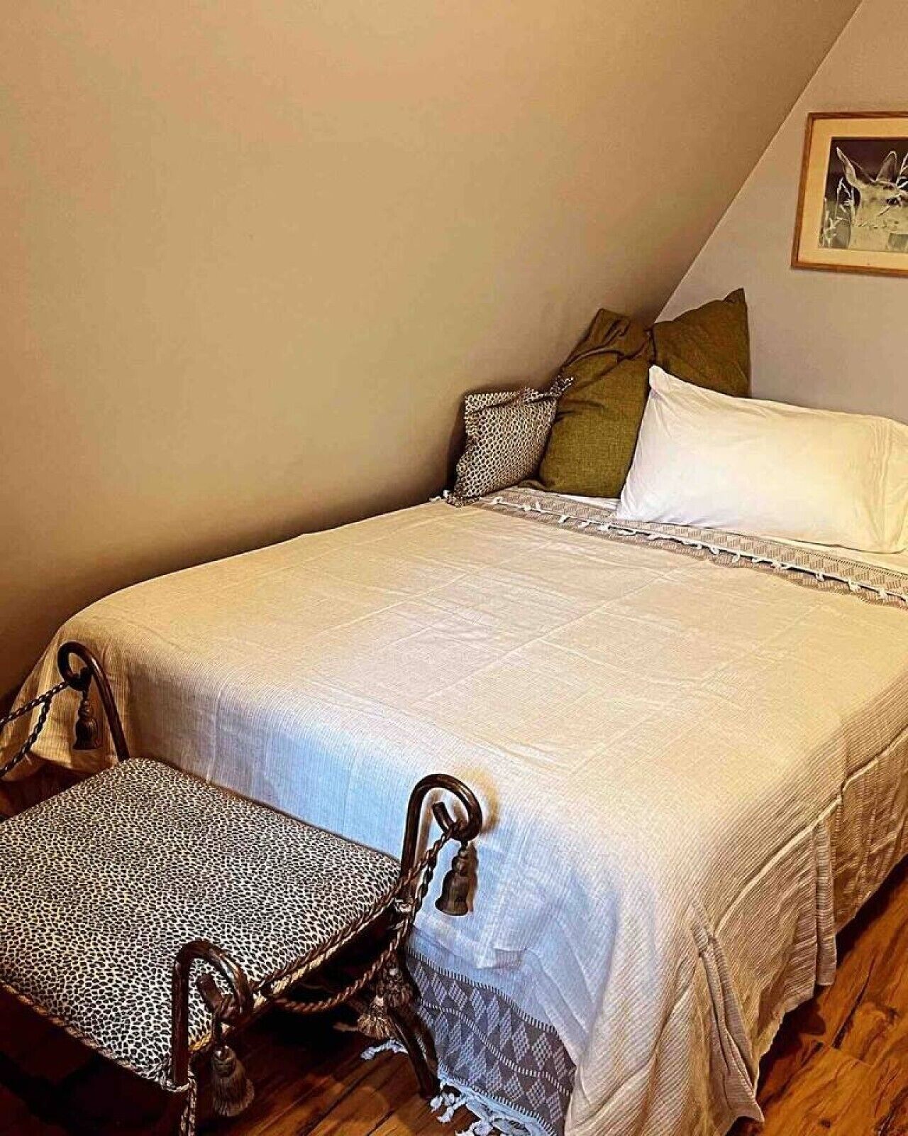 A single bed with a white bedspread and a matching pillow, a wooden chair with a leopard print cushion, and a framed picture on the wall.