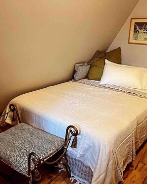 A single bed with a white bedspread and a matching pillow, a wooden chair with a leopard print cushion, and a framed picture on the wall.