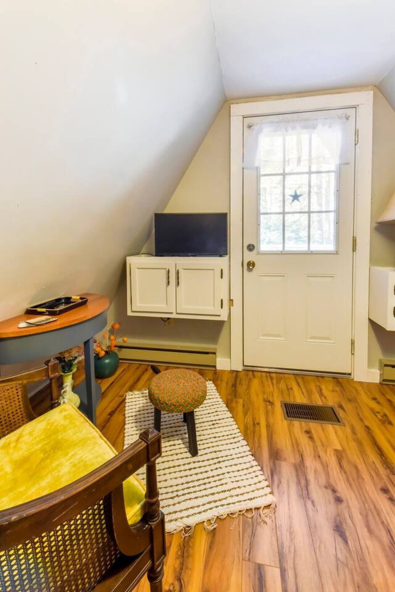 Una habitación con techo inclinado, una puerta blanca decorada con una estrella, una pequeña mesa redonda con una silla y un televisor sobre un soporte.
