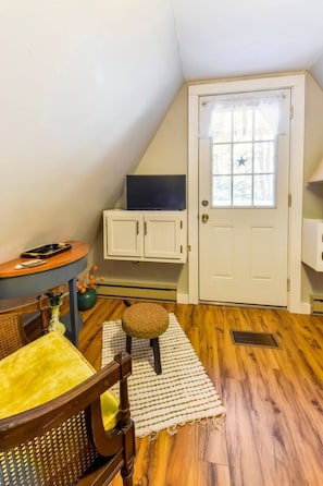 A room with a sloped ceiling, a white door with a star decoration, a small round table with a chair, and a television on a stand.
