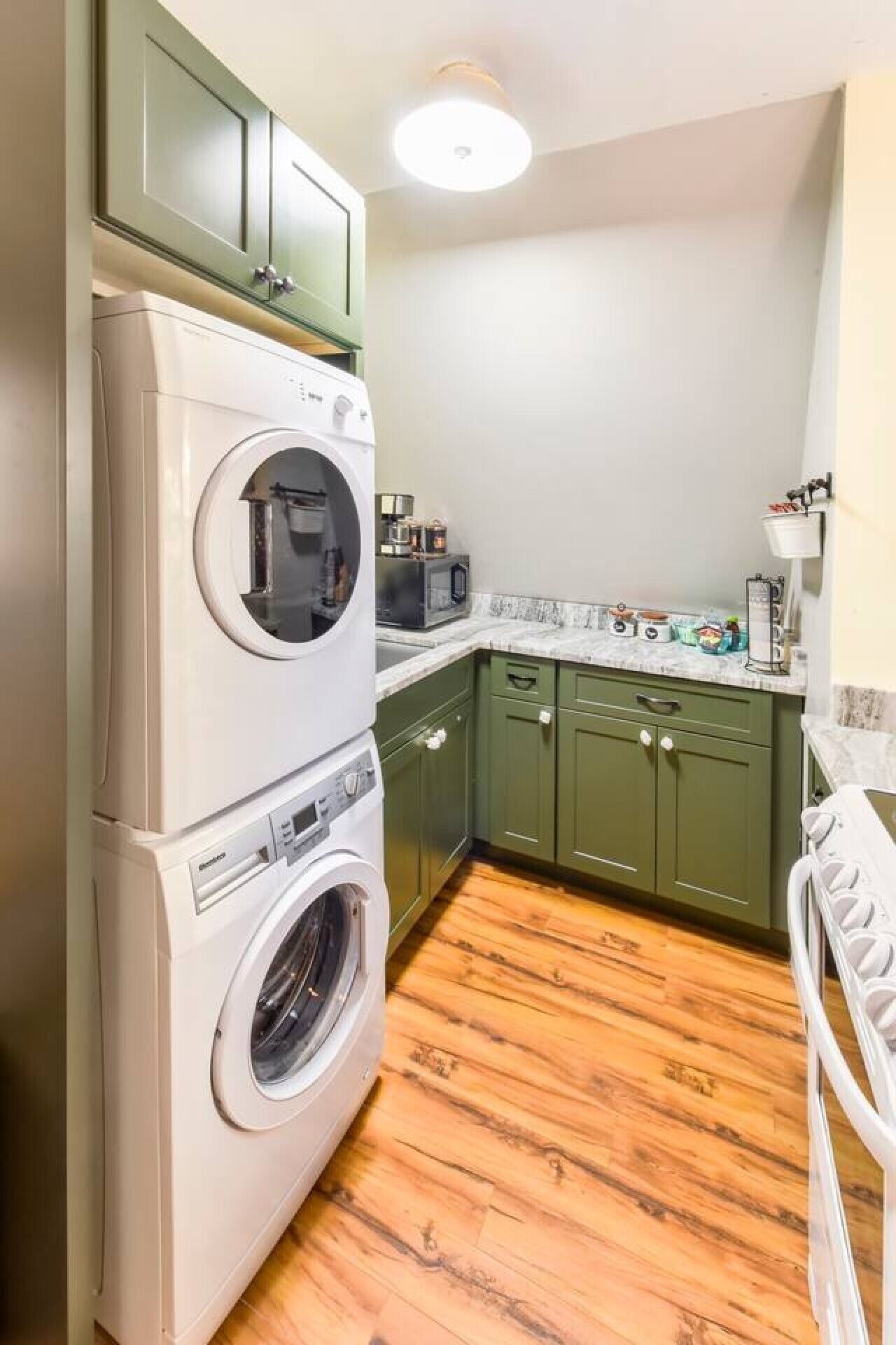 A compact kitchen with a stacked washer and dryer, green cabinets, a countertop with various appliances, and a wooden floor.