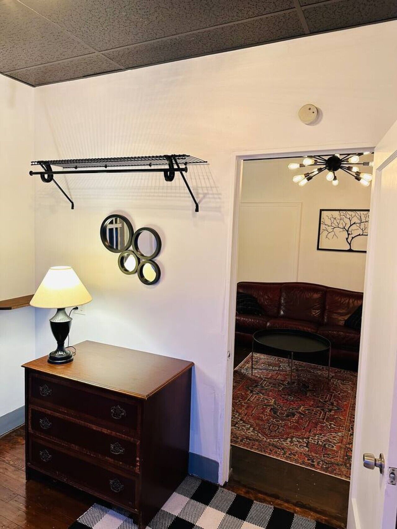 Een kamer met een houten dressoir, een lamp, een droogrek en een deur naar een woonruimte met een bank en een kamerbreed tapijt.