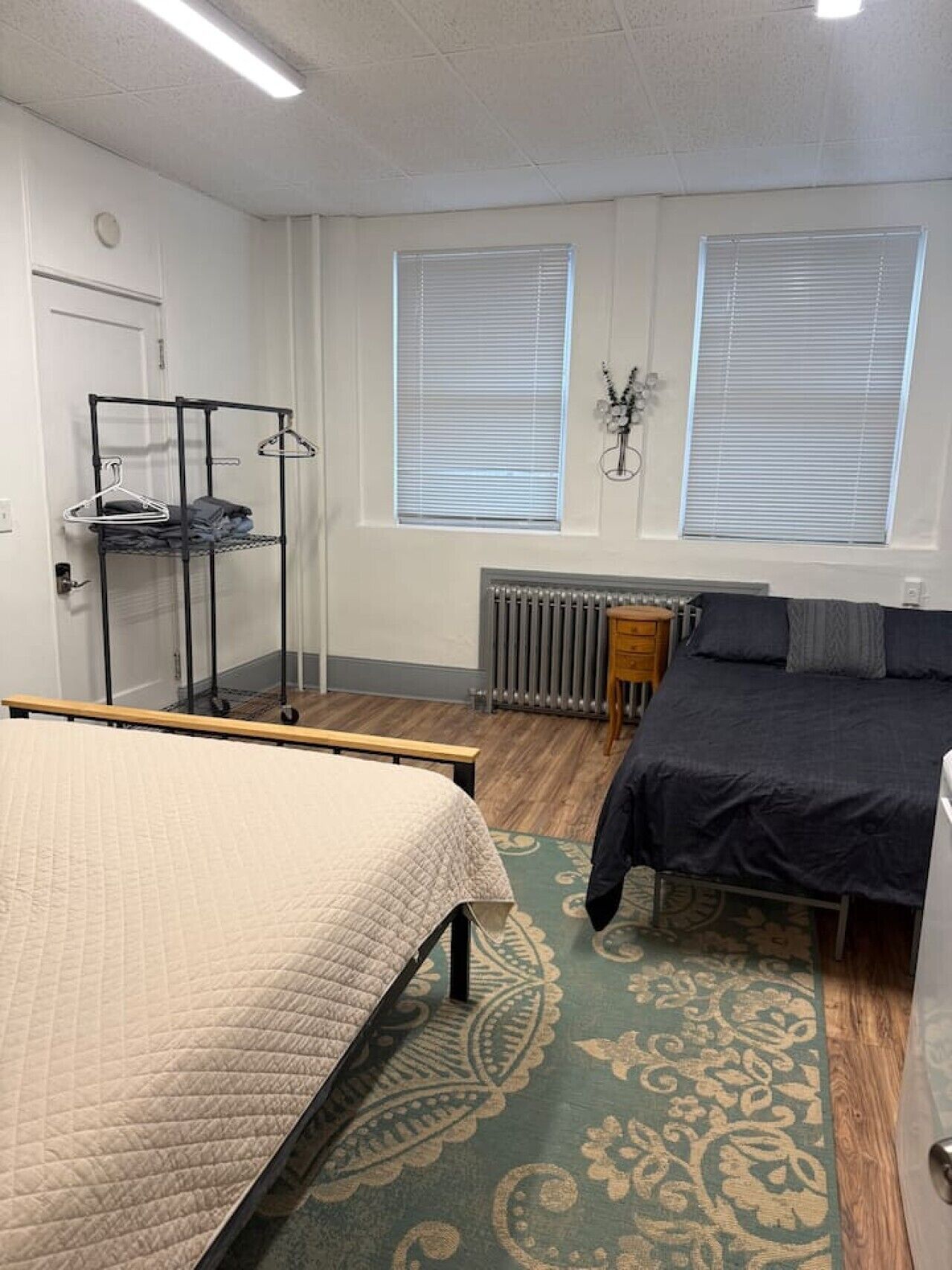 A bedroom with a bed, a dresser, a radiator, a shelf with clothes, and two windows with blinds.