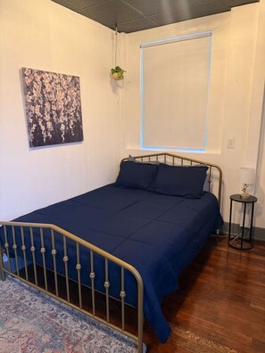 Un dormitorio con una cama, una mesita de noche pequeña, una planta colgante y un cuadro en la pared.
