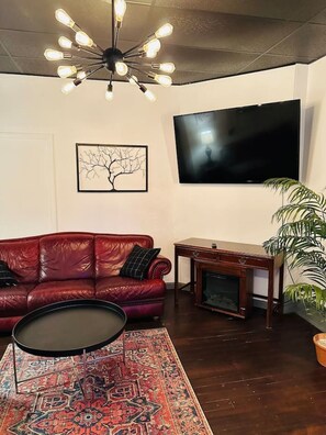Una sala de estar con un sofá de cuero rojo, un televisor de pantalla plana, una mesa de centro, una alfombra estampada y una planta en maceta.