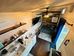A compact kitchen with a sink, stove, and countertop. There is a small seating area with a cushion and a window with curtains.