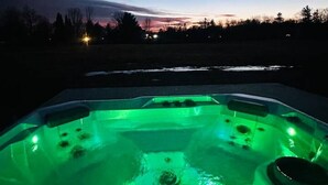 Un bain à remous éclairé de lumières vertes, entouré d’un paysage naturel et surplombant un ciel crépusculaire.