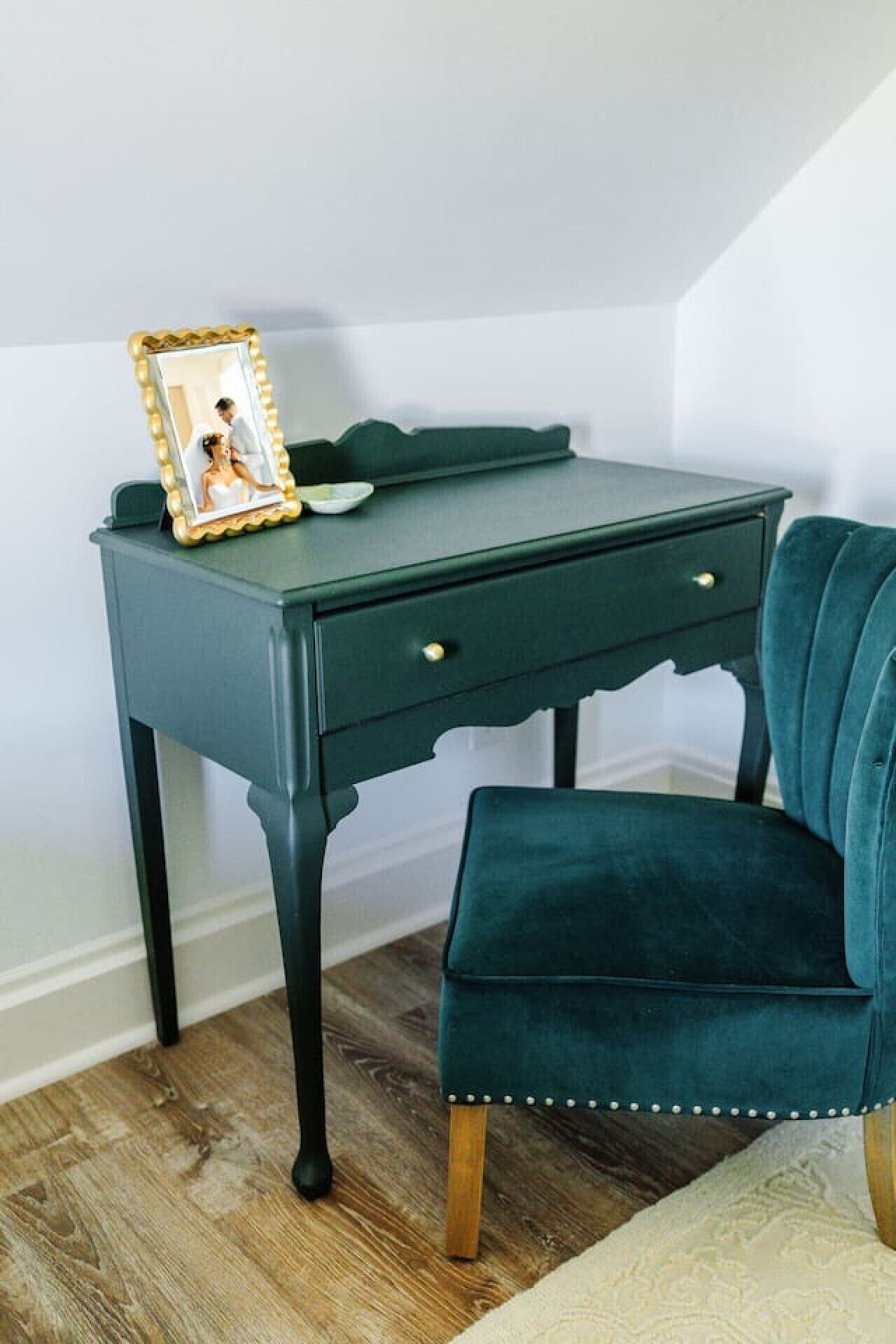 A dark green desk with a picture frame and a small bowl, a teal upholstered chair with wooden legs, and a wooden floor.