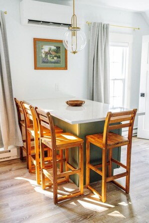 Een eetgedeelte met een wit eilandtafeltje, omringd door houten stoelen, een hanglamp en een gekaderd schilderij aan de muur.