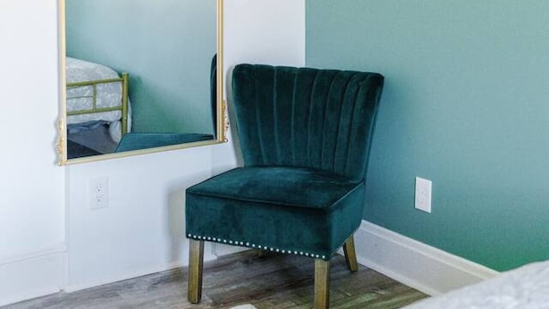 A room with teal walls, a dark green velvet chair, a large mirror with gold trim, and framed artwork.