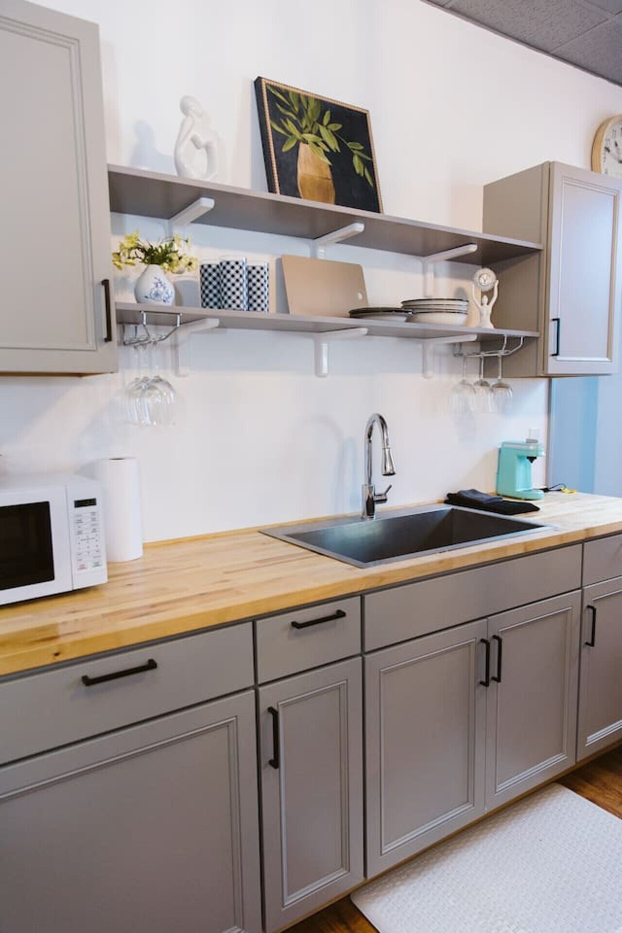 A modern kitchen with grey cabinets, a wooden countertop, a sink, a microwave, and a clock on the wall.