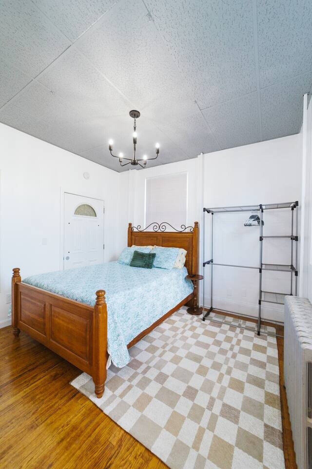 Ein Schlafzimmer mit einem hölzernen Bett, einem karierten Teppich, einem Wäscheständer aus Metall und einer weißen Tür.