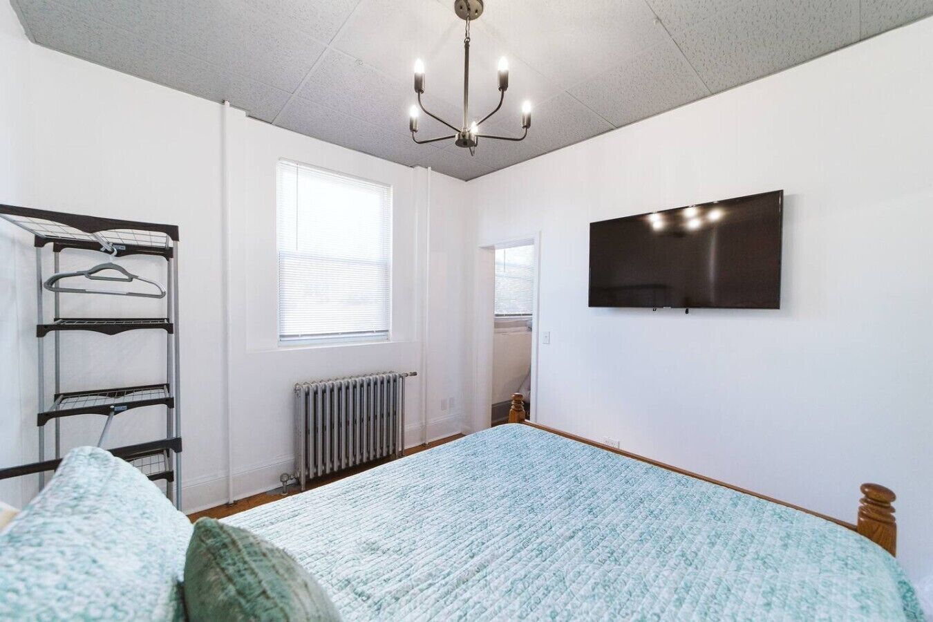 A bedroom with a bed, a radiator, a wall-mounted TV, and a window with blinds.