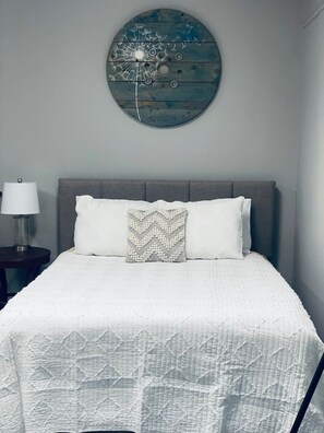 Een slaapkamer met een bed, nachtkastje, lamp en een kunstwerk aan de muur.