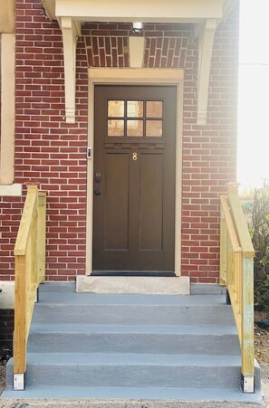 A brown front door with a window above it, a brick wall, and a set of wooden stairs leading up to the door.