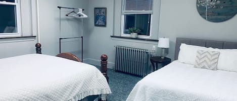 A hotel room with two beds, a radiator, a window with blinds, a hanging light fixture, and a wall clock.