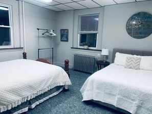A hotel room with two beds, a radiator, a window with blinds, a hanging light fixture, and a wall clock.