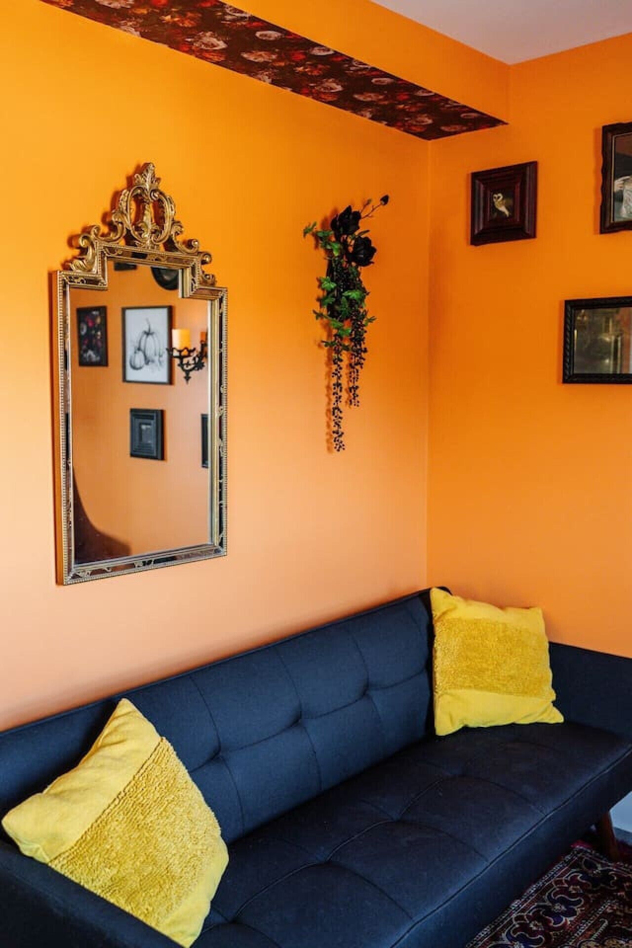 A room with a dark blue sofa, two yellow cushions, a large mirror with ornate gold detailing, and a floral hanging decoration.