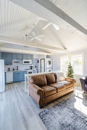 A spacious living area with a brown leather sofa, a kitchen with blue cabinets, and a ceiling fan.