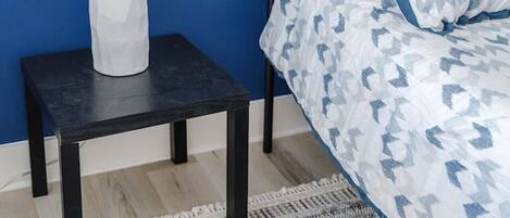 A bed with a patterned bedspread and pillows, a bedside table with a lamp, and a dark floor.