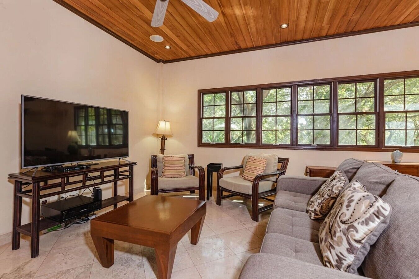 A living room with a television, wooden furniture, a sofa, and large windows.