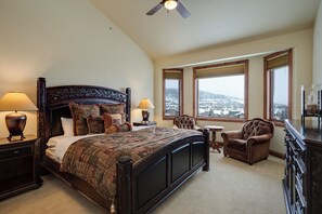 A bedroom with a large bed, two bedside lamps, a chair, a small table, and a view of a snowy landscape through the windows. - AC1465 | Perfect Mtn Getaway | Beautiful Valley Views | Close to slopes (Steamboat Springs)