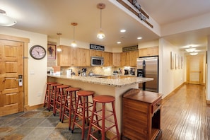 A kitchen with wooden cabinets, a stainless steel refrigerator, a microwave, and a bar area with stools. - AL4102 | Perfect Mountain Home | 10 Hot Tubs | Air Chilling | Pools (Steamboat Springs)