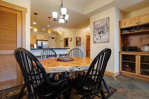 A dining area with a wooden table and chairs, a chandelier, and a kitchen in the background. - AL4109 | Private Courtyard with Hot Tub | Pools | Close to Base | Air Chilling (Steamboat Springs)
