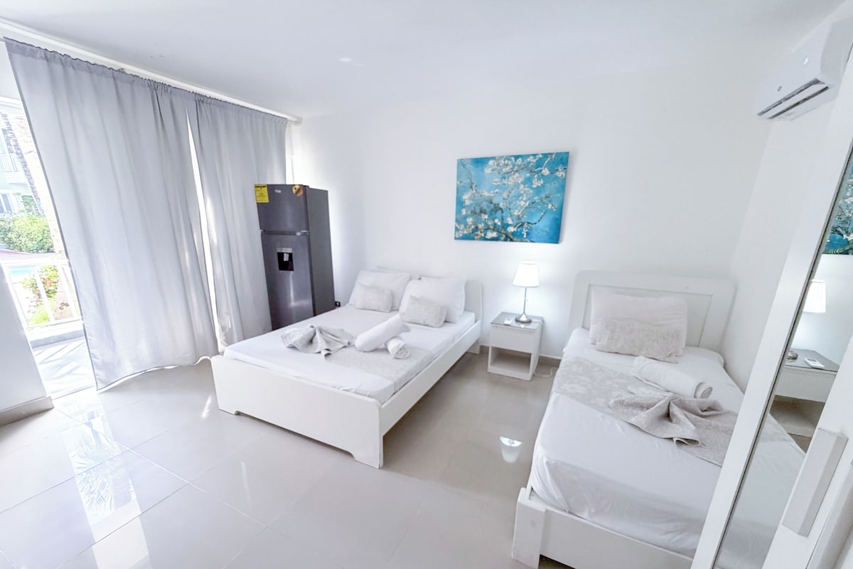 A hotel room with two beds, a refrigerator, a painting, and a view of the outdoors.