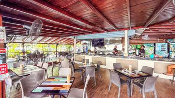 An outdoor restaurant with wooden ceiling beams, wicker chairs, and a bar area with a chef preparing food.