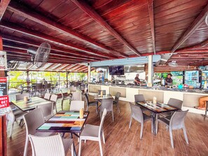 Un restaurant en plein air avec des poutres en bois au plafond, des chaises en osier et un bar où un chef prépare les plats.
