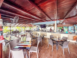An outdoor restaurant with wooden ceiling beams, wicker chairs, and a bar area with a chef preparing food.