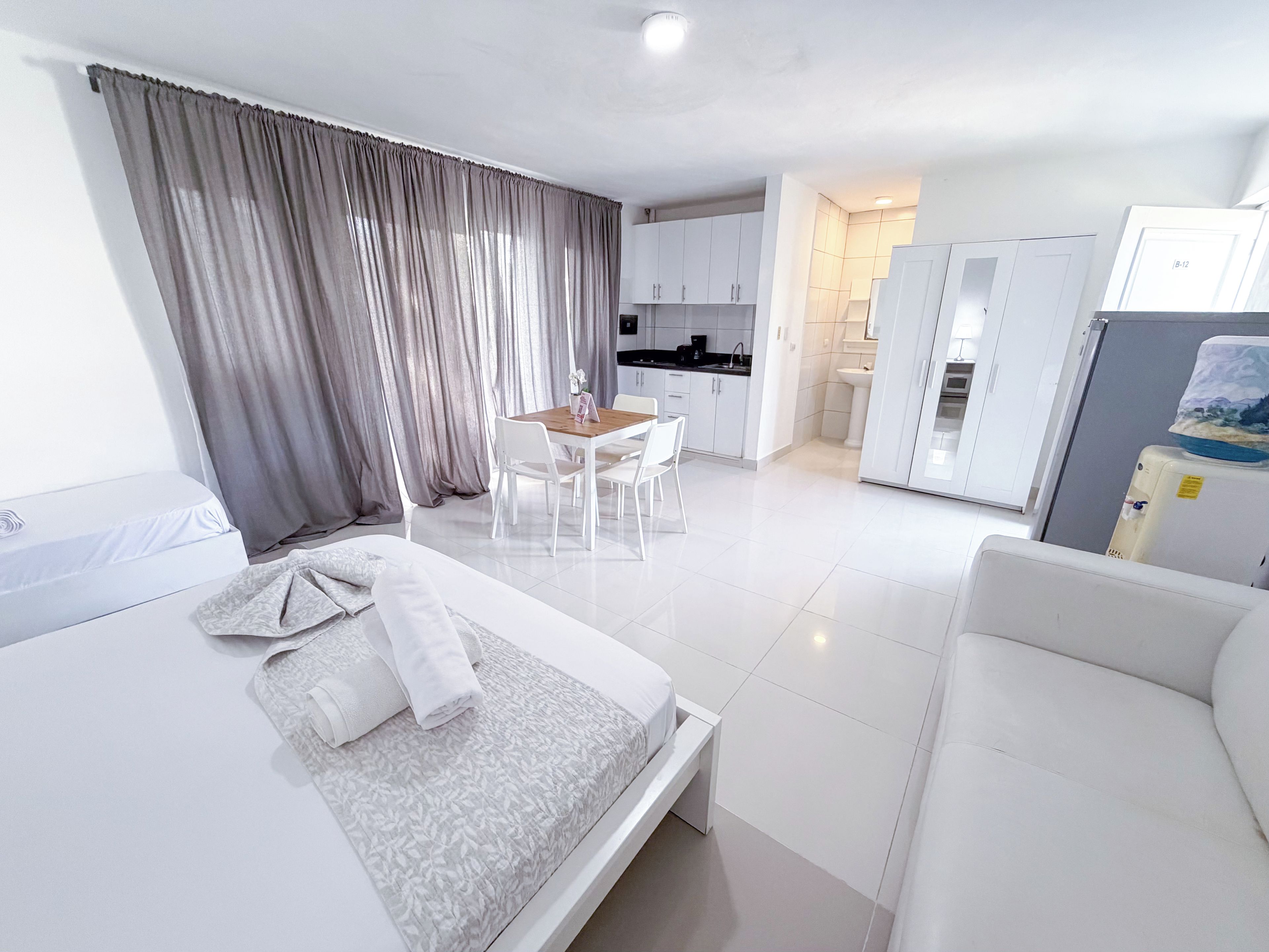 Una habitación de hotel moderna con cama, una pequeña mesa de comedor con sillas, kitchenette y baño con lavamanos e inodoro.