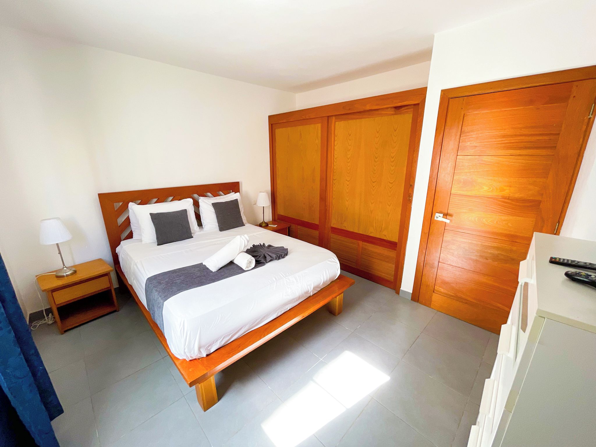 Habitación de hotel con cama, mesita de noche, lámpara, armario y escritorio con control remoto.