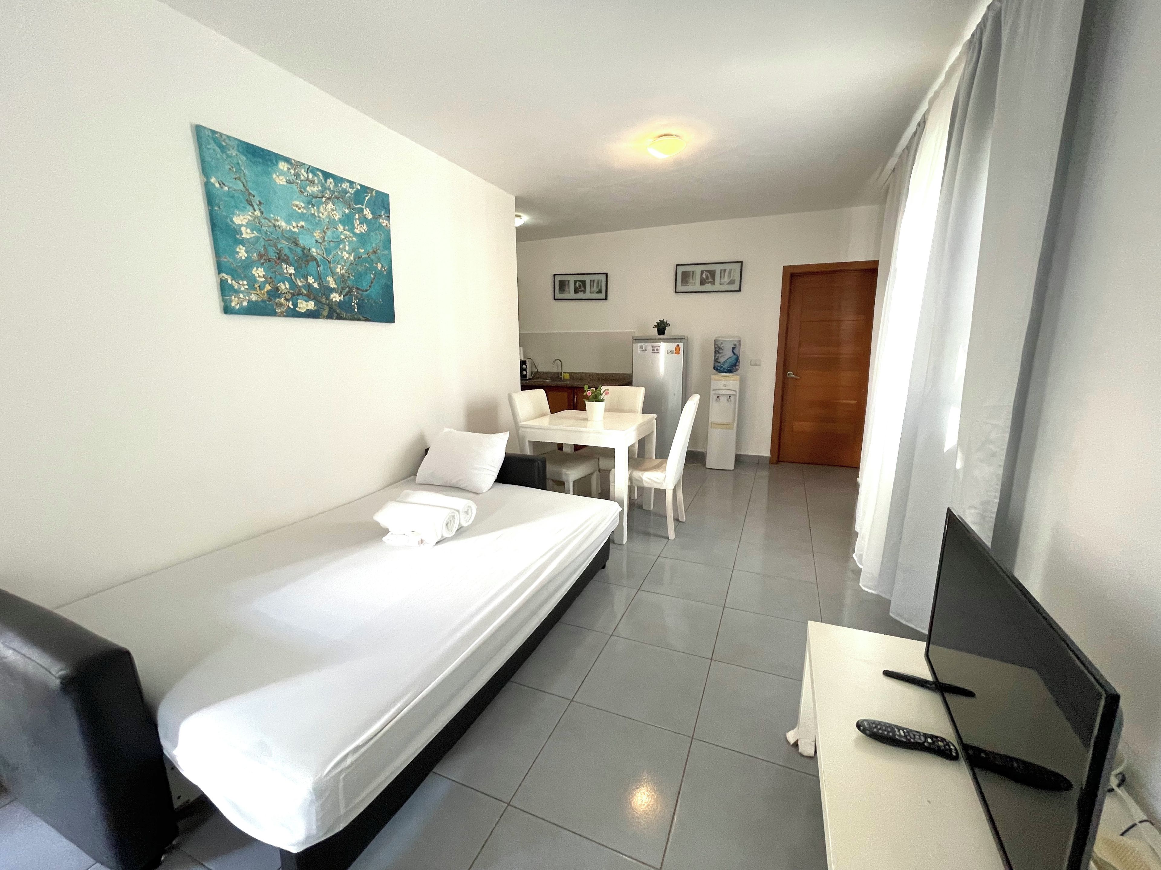 Ein Hotelzimmer mit Bett, Schreibtisch mit Computer und Fernbedienung, Esstisch mit Stühlen, einer Küchenzeile und einem Bild an der Wand.