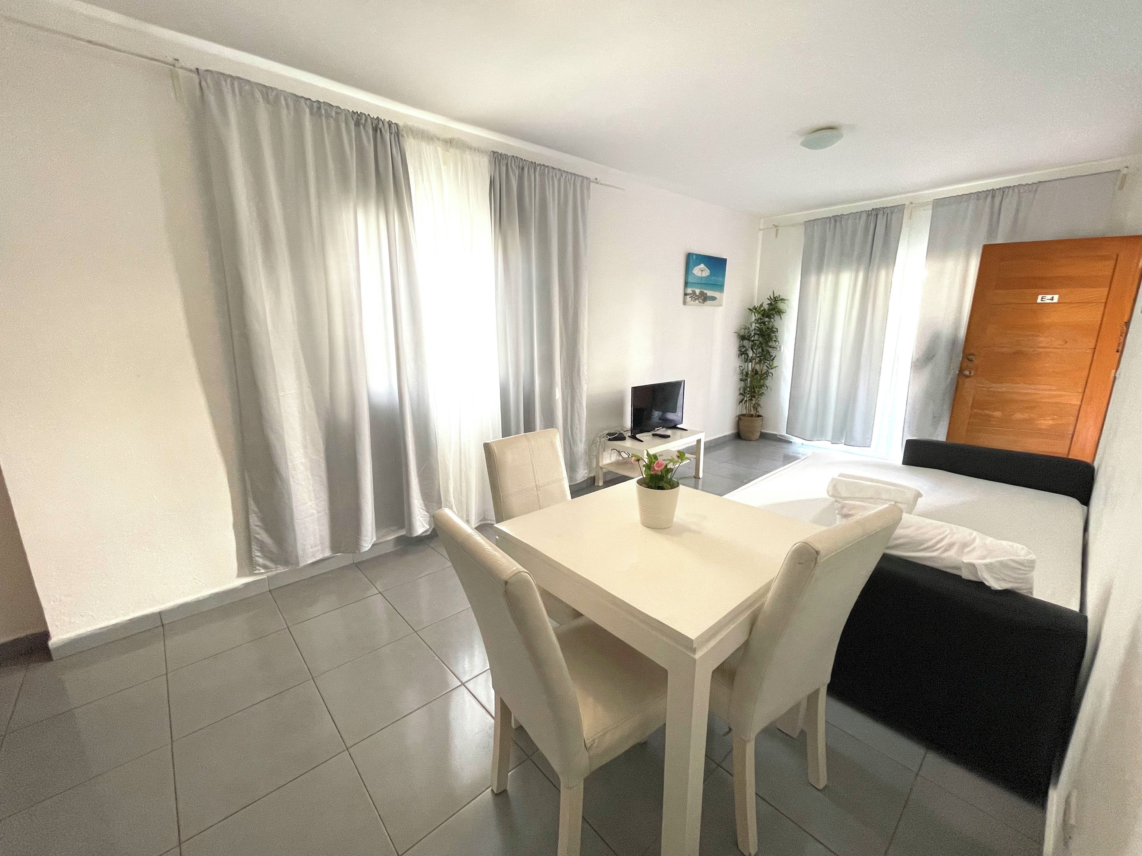 A hotel room with a bed, a TV, a dining table with chairs, a potted plant, and a window with curtains.