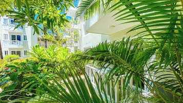 Un exuberante jardín con palmeras y diversas plantas, un edificio blanco con balcones y un cielo azul despejado.