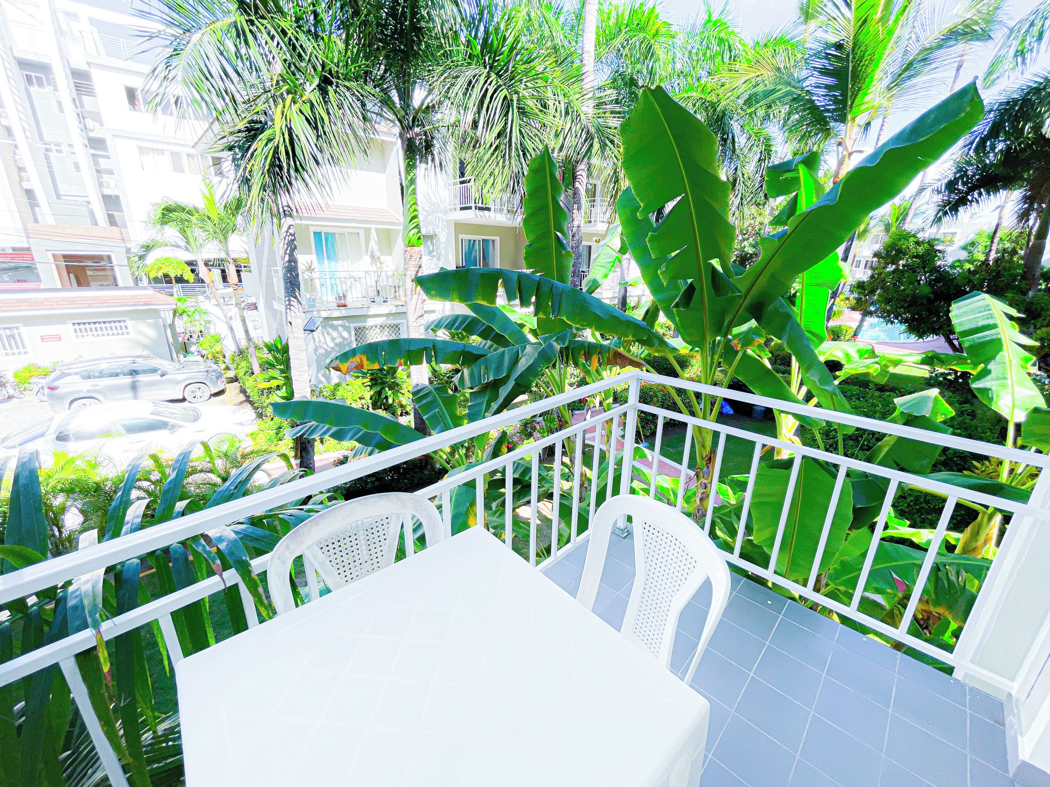A balcony with white chairs and a table, surrounded by lush green plants and a view of a residential area.