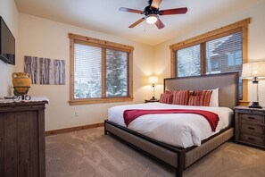 Une chambre à coucher comprenant un lit, deux tables de chevet, un ventilateur de plafond et de grandes fenêtres équipées de stores.