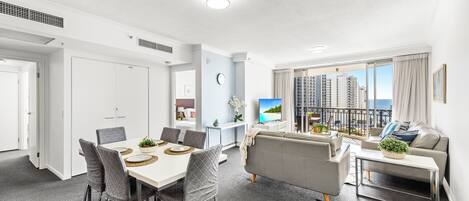 A modern living room with a grey sofa, a dining table with chairs, a TV, and a balcony with a city view.